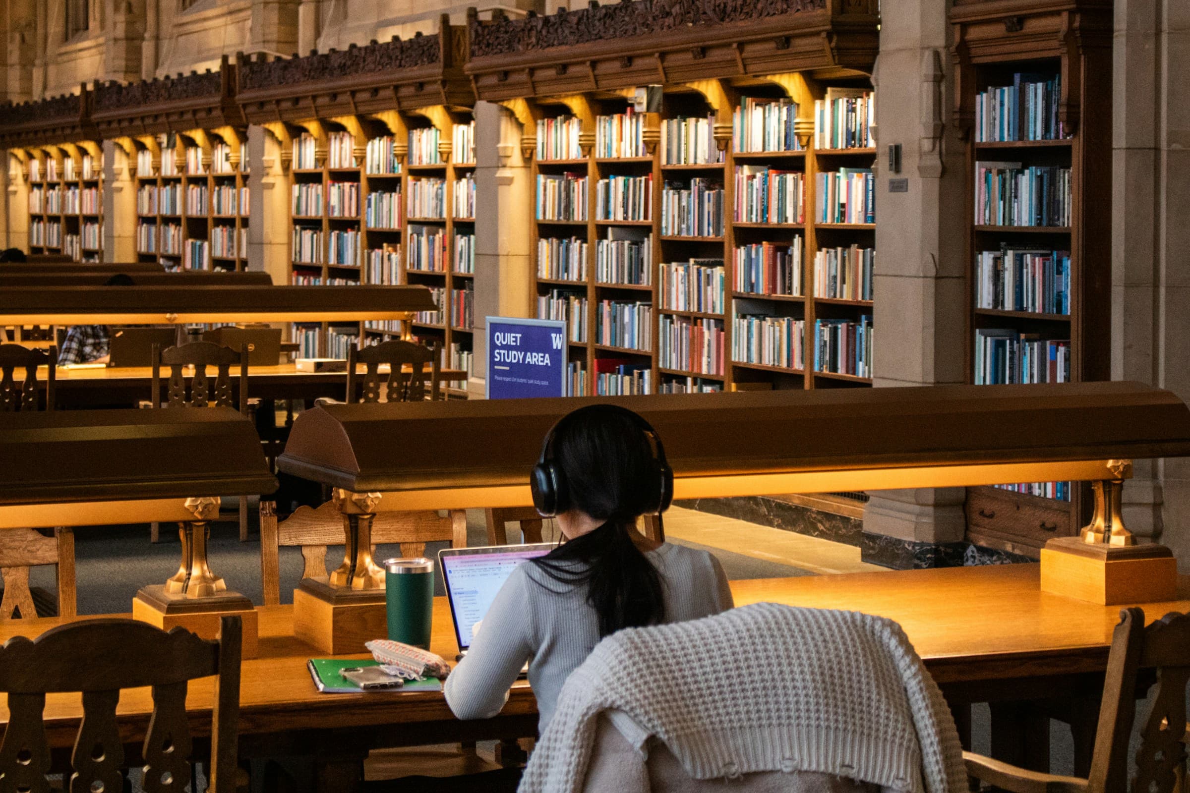 A student with headphones studying at a lamp-lit desk in a library